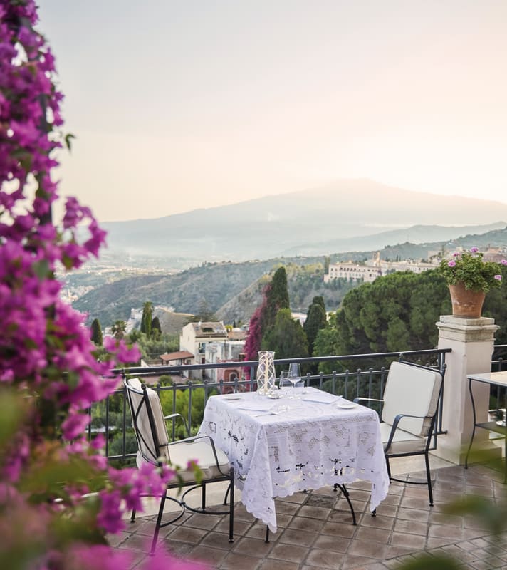 A table for two with a lace cloth overlooks Taormina's romantic hills from the terrace edge, seen through pink bougainvillea.