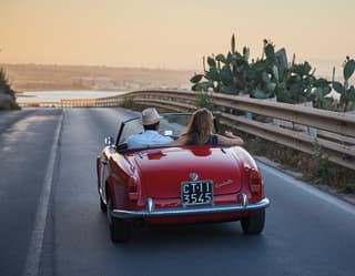 Gleaming, red, soft-top classic car on a seaside road at sunset