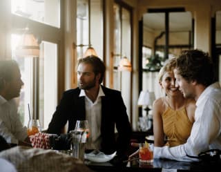 A woman in a yellow dress and three men in smart shirts converse and drink orange-garnished Aperol spritz at Bar Timeo.