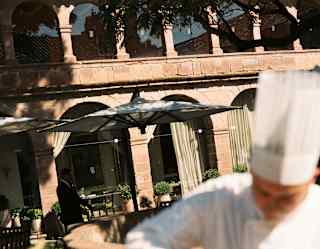 A chef in soft-focus foreground, in whites and a toque, prepares dishes in the courtyard, seen in bright sunshine.