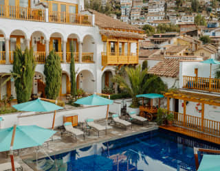 High-angle view over a blue pool, flanked by parasols, loungers and hotel walls, with a glimpse of hillside dwellings behind.