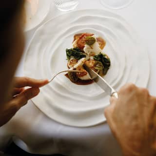 Close-up of two hands holding a knife and fork and cutting into a fig dessert on a white plate