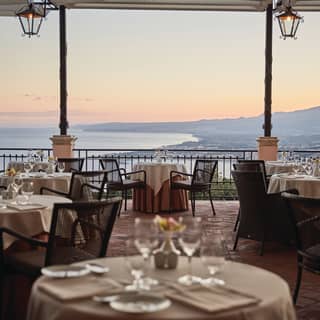Formal linen-coated table in the foreground of a restaurant terrace with views of Taormina at dusk beyond