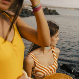 Two women in swimsuits stand on rocks at the water's edge. One clutches a straw hat as she gazes out over the Mediterranean