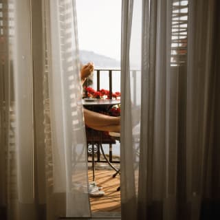 View from a shadowy suite through diaphanous drapes onto the balcony where a guest sits with feet up, enjoying the views.