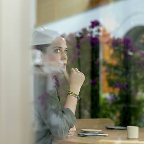 Chef Pía León looks pensive as she sits at a table, viewed from the outside through a window that reflects strips of sky.