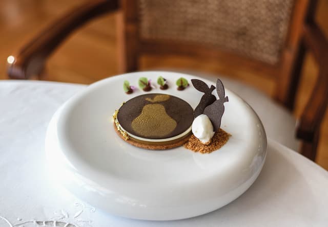A pear, ricotta and caramel dish with a chocolate-topped disc with gold motif and two pear-shaped wafers on cream and crumbs.